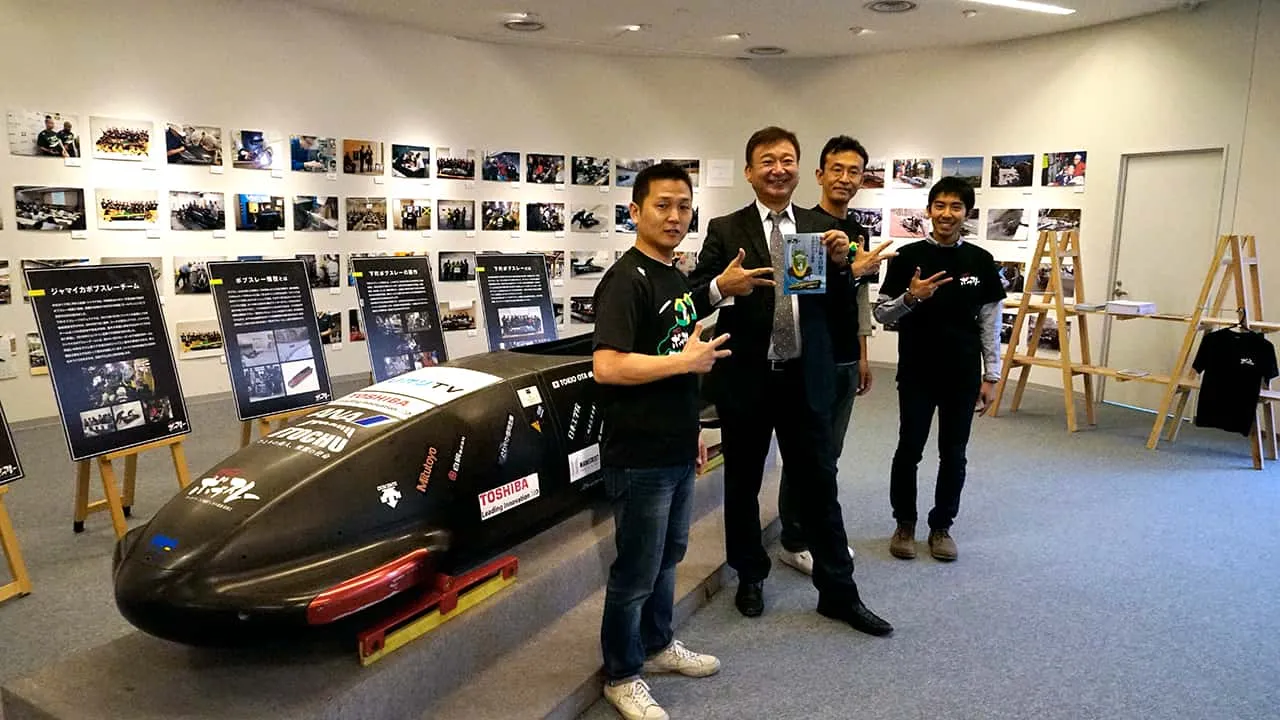 スポンサーロゴが並ぶ黒い実機の前で、お揃いのポーズで記念撮影