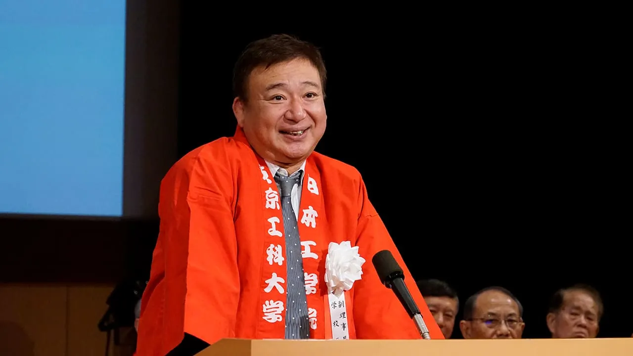 襟に日本工学院、東京工科大学と入った法被に白い花の胸章をつけ、笑顔で挨拶する校長
