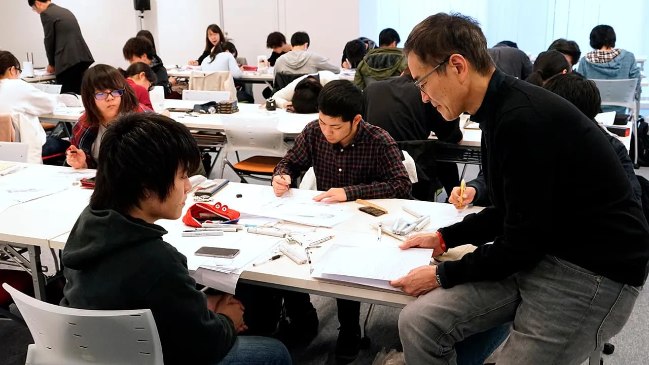 並べられたテーブルについてデザイン制作をする学生達を見回りながら、随時指導を行っている様子