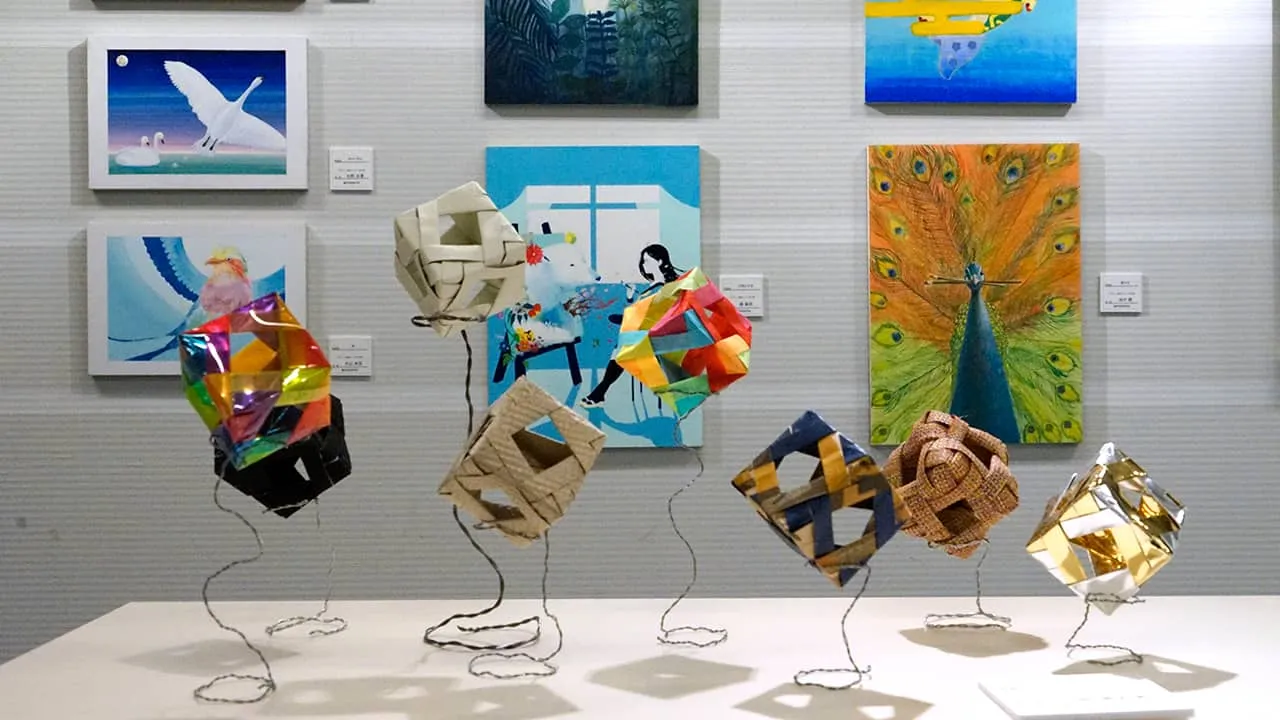 絵画系の壁面展示の手前に、様々な色や材質の立方体を針金のスタンドで浮いているように展示した作品の写真