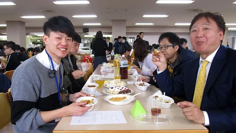 着席式の会場にて、留学生と対面で座りカレーを食べながら1枚。口の中がいっぱいの校長。