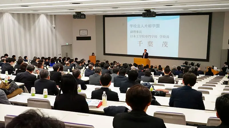 壇上に校長が立っている風景を階段状の座席後方から撮影した写真