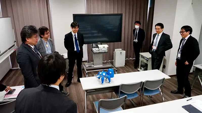 とあるアニメのロボットに見えるAIロボットを囲み、楽しそうなスーツ姿の大人たち