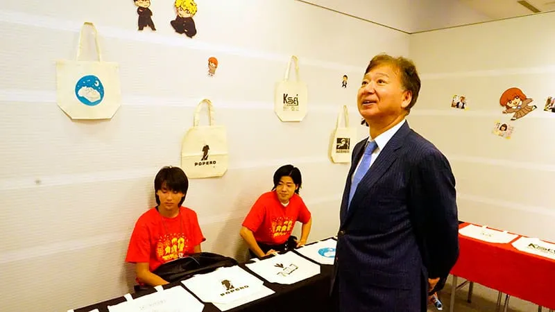 展示物を見上げる千葉理事長とその背後で緊張の面持ちなスタッフ（生徒）2人