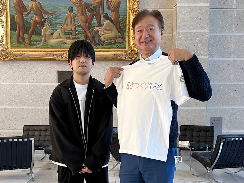 お気に入りの作品を体の前で広げる千葉理事長と、作品を作成した男子学生のツーショット