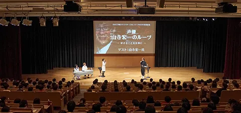 壇上に立ち向かい合ってトークする2人とそれを聞く生徒たち