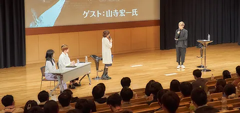 壇上に立ち向かい合ってトークする2人とそれを聞く生徒たち