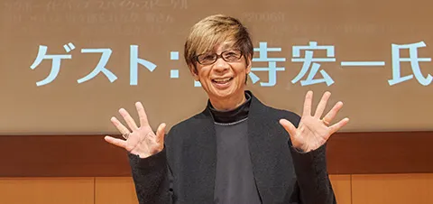 顔の左右斜め下で両手を開くポーズで笑顔の山寺さん