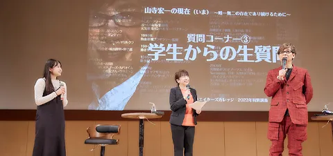 壇上でトークをする山寺さんをその横で見つめる富永先生と女子生徒