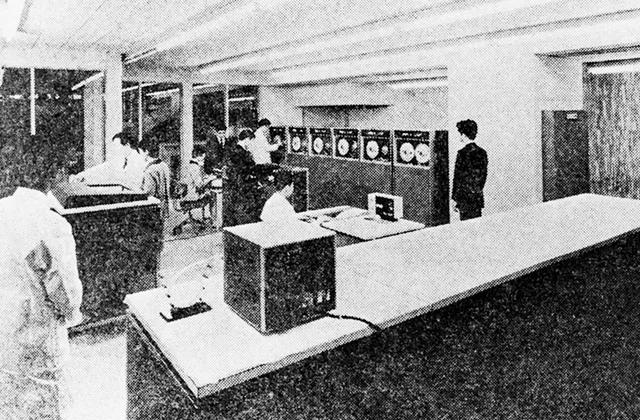 成人男性の身長ほどもある大型電子計算機が何台も設置されている室内で実習を行っている白黒写真