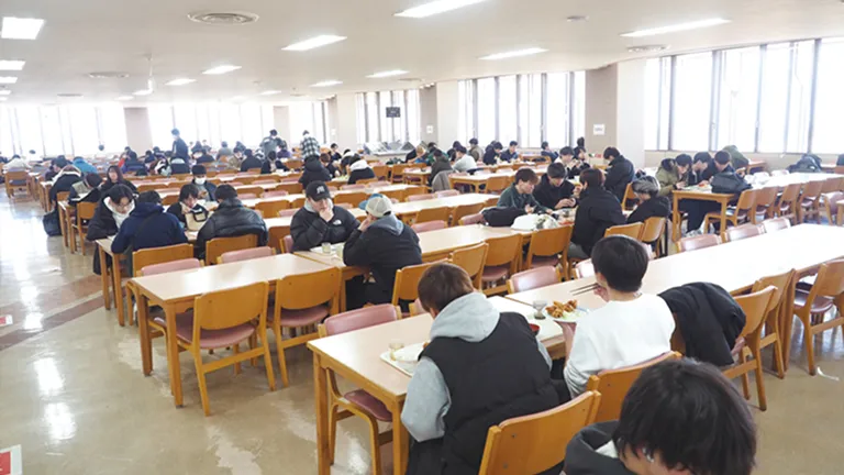 窓に囲まれたとても広い空間にしっかりした木製のテーブルと椅子が並んだ食堂で生徒達が食事をしている画像