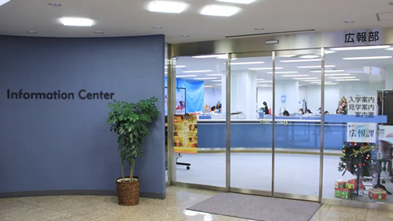 弧を描いた落ち着いた色調の青い壁にインフォメーションセンター、ガラス張りの入り口の枠に広報部の表記あり、観葉植物や季節の装飾品が飾られている風景