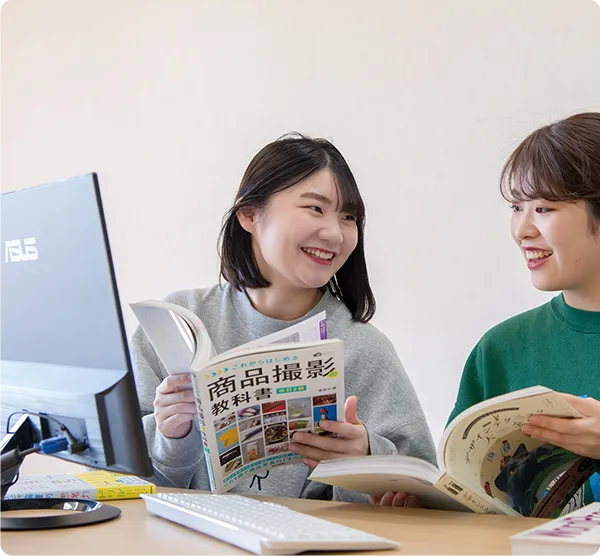 パソコンと賞品撮影の教本を見ながら会話する二名の学生
