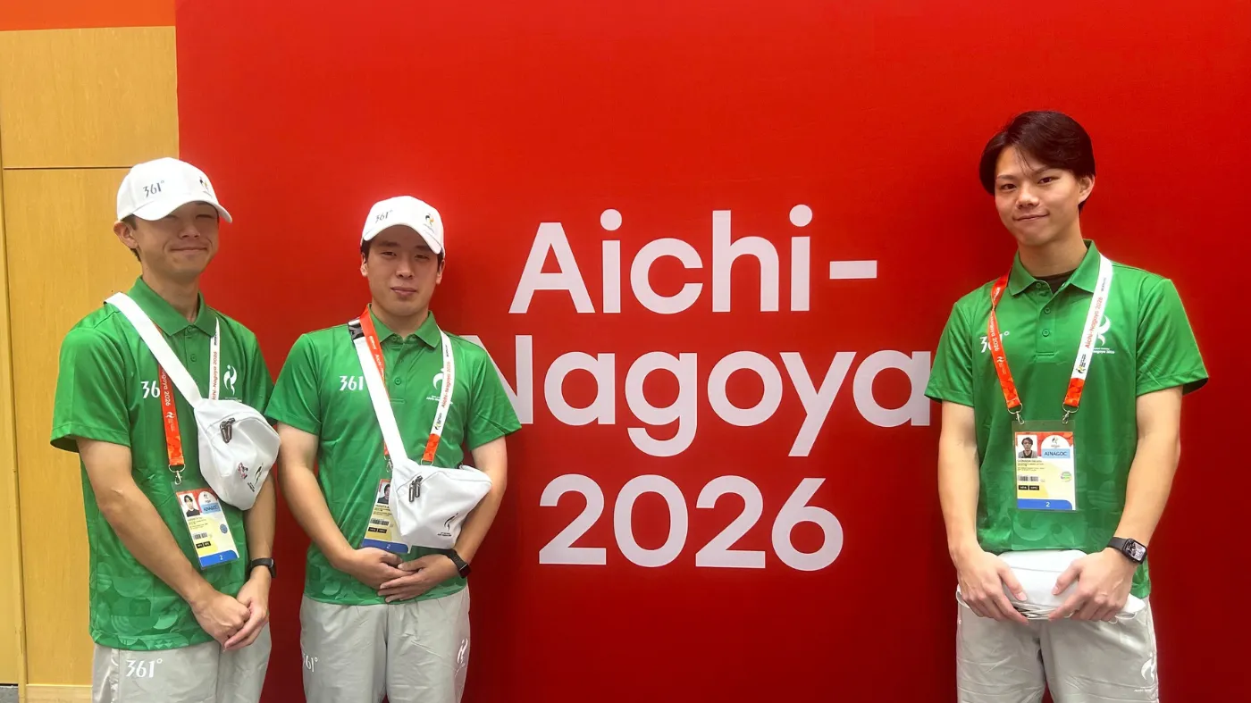 「Aichi-Nagoya 2026」のサイン前で記念撮影する大会スタッフ3人。