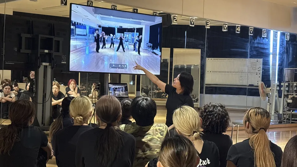 モニターに映るダンス映像を指しながら解説する講師とそれを聞く学生たち
