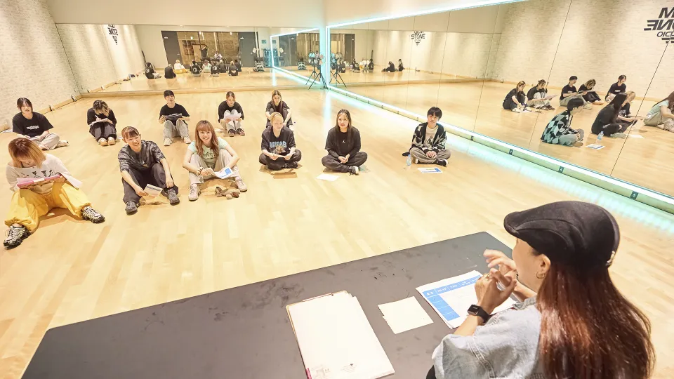 講師の話をレッスン場の床に座り笑顔で聞く学生たち