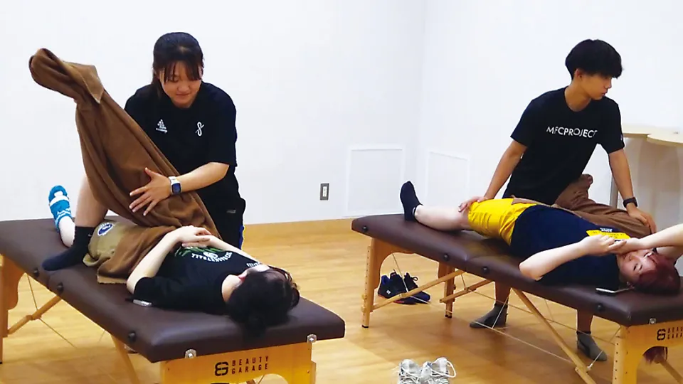マッサージ台の上に横たわる学生が、トレーナーから足のストレッチを受ける様子